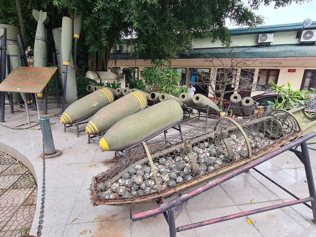 A display of various types of military ordnance, including bombs and shells, arranged on a concrete surface with greenery in the background.