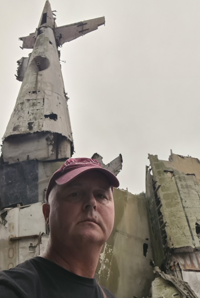 A person in a cap poses for a selfie in front of a large, weathered airplane wreckage with a damaged vertical stabilizer, set against a cloudy sky.