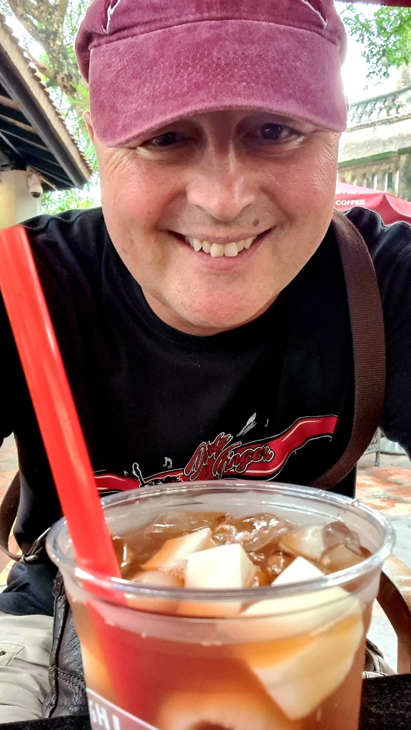 A smiling person wearing a maroon cap and a black t-shirt with a design, sitting at a table with a drink that has ice and pieces of fruit, surrounded by outdoor seating and greenery.