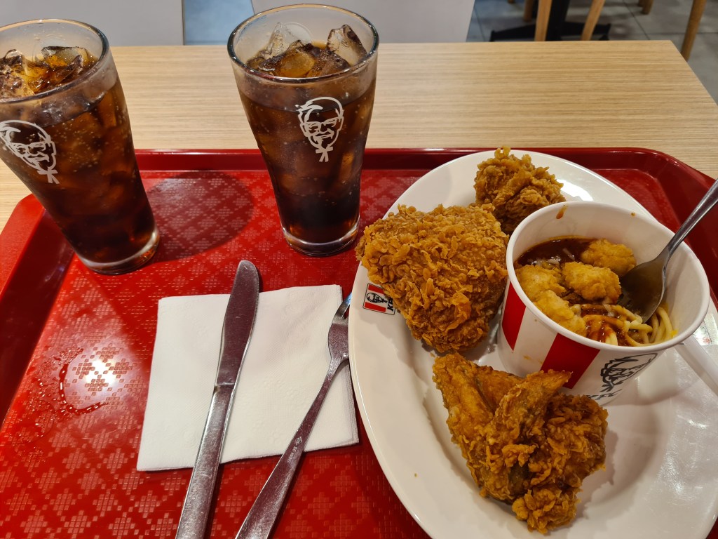 A tray featuring two glasses of iced cola, a plate with three pieces of fried chicken, and a cup of noodles with sauce and breaded chicken pieces.