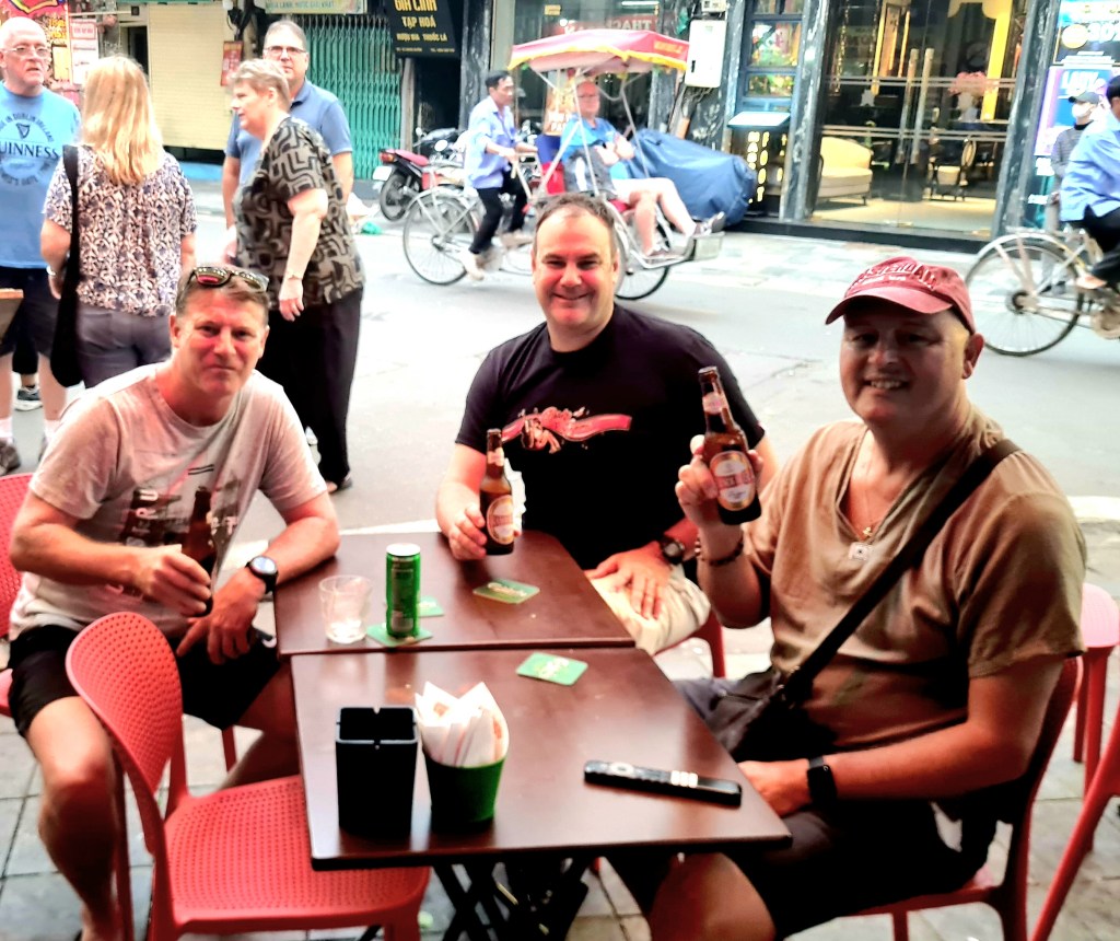 Three men sitting at a table in a busy street, each holding a beer, enjoying their time together. The background shows pedestrians and a bicycle rickshaw.