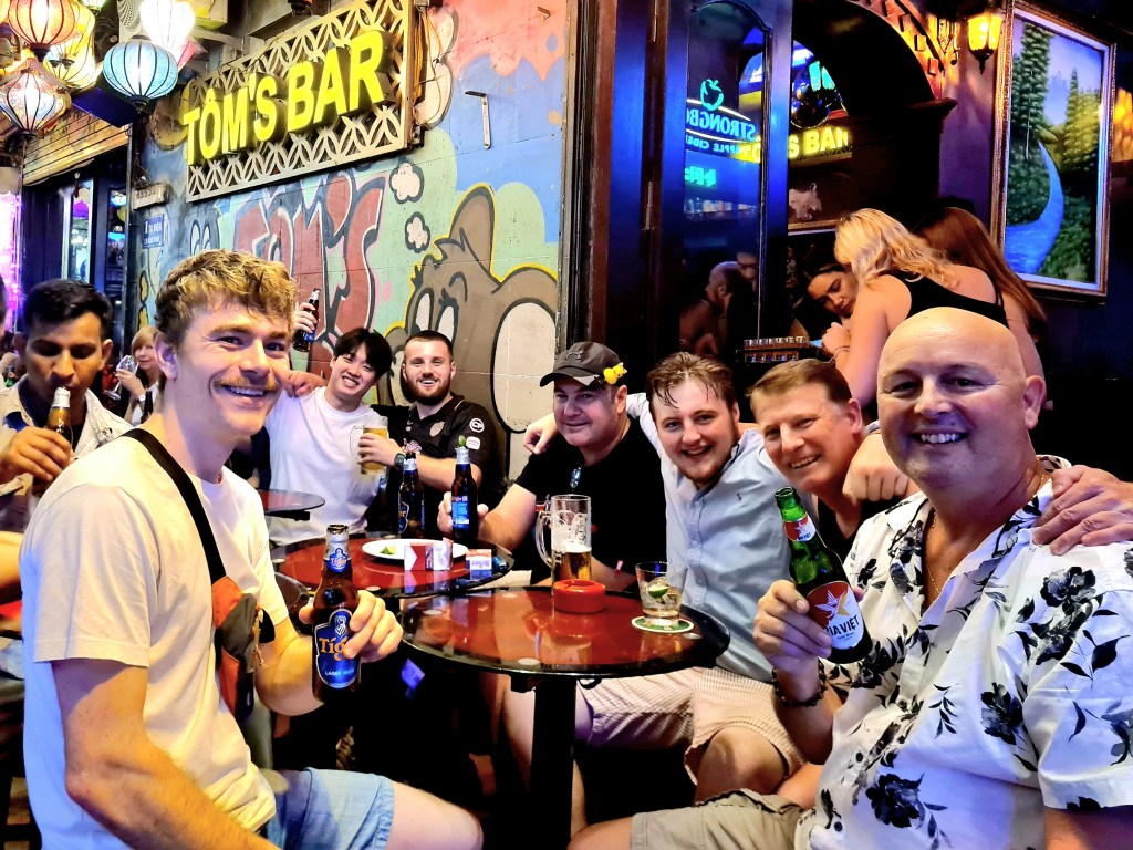 A group of seven men sitting around a table at Tom's Bar, enjoying drinks and smiling at the camera. The bar features colorful lanterns and vibrant wall art.