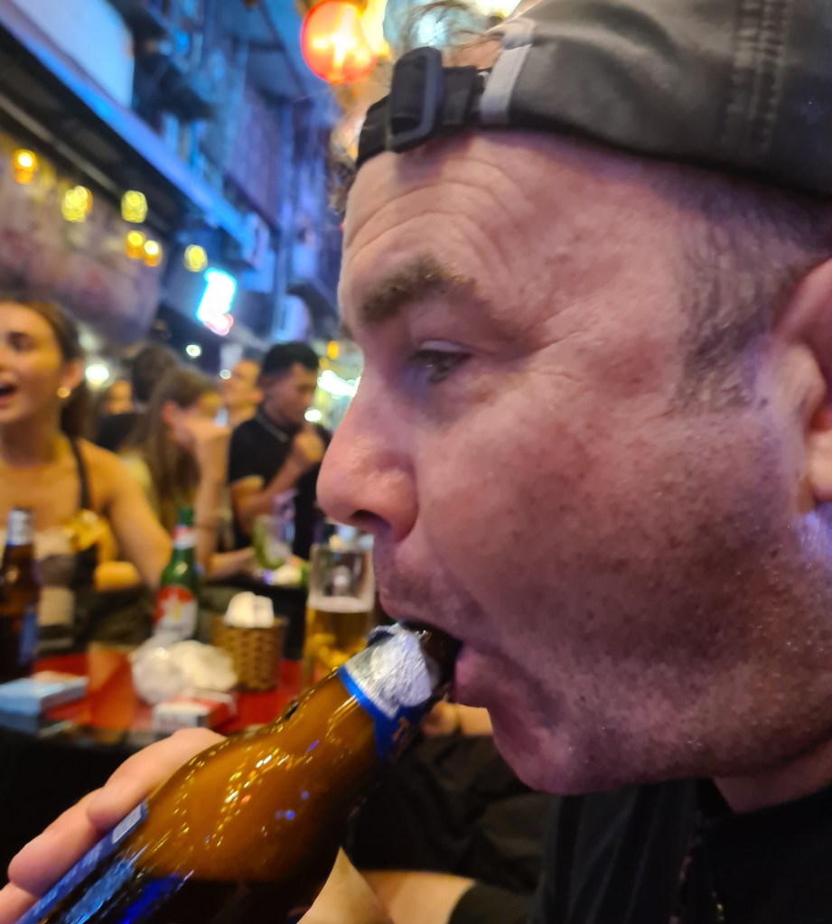 Close-up of a man taking a drink from a bottle in a lively bar setting, with people and colorful lights in the background.