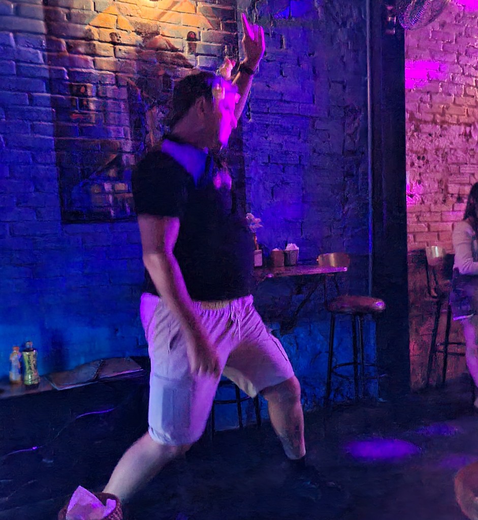 A person dancing energetically in a dimly lit venue with colorful lights and a brick wall background.