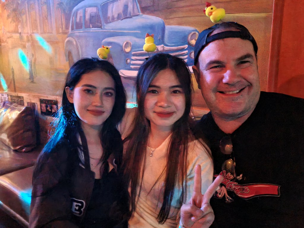 Three people smiling together, with two women on the left and right wearing casual outfits, and a man in the middle wearing a black cap. They all have small rubber duck toys on their heads, posing in a colorful setting with a vintage car mural in the background.