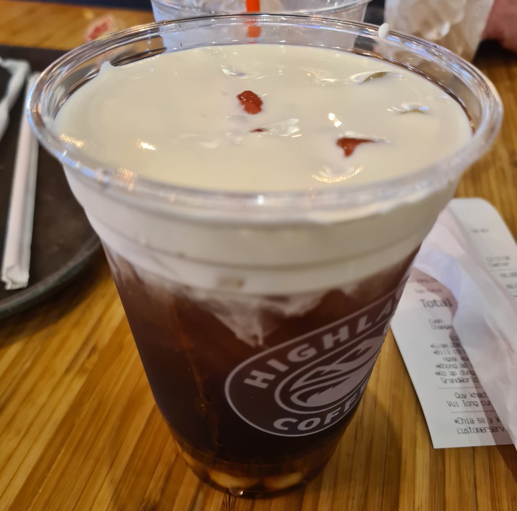 A cup of iced coffee topped with creamy foam, featuring the logo of Highland Coffee, placed on a wooden table.