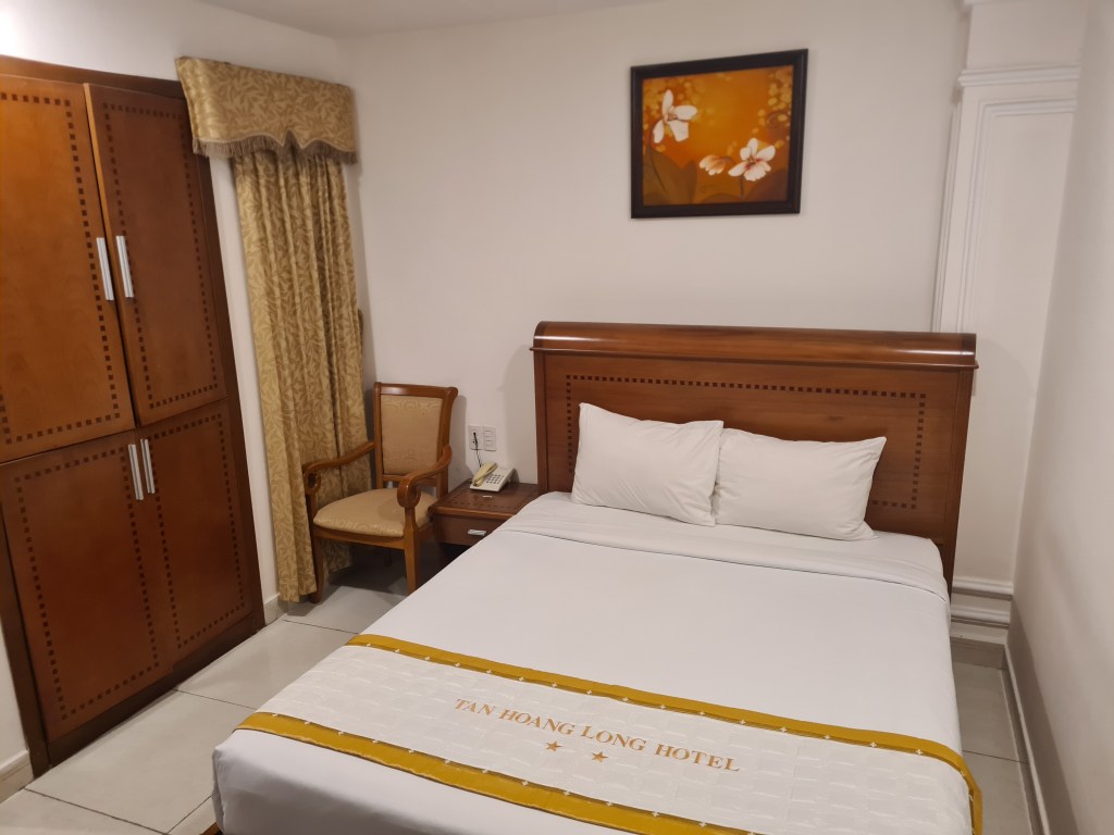 A cozy hotel room featuring a single bed with white linens, a wooden headboard, and decorative gold trim. There's a small bedside table with a telephone, a chair beside it, and a wardrobe with a smooth wooden finish. The wall displays a framed painting of flowers, and there are curtains that complement the room's decor.