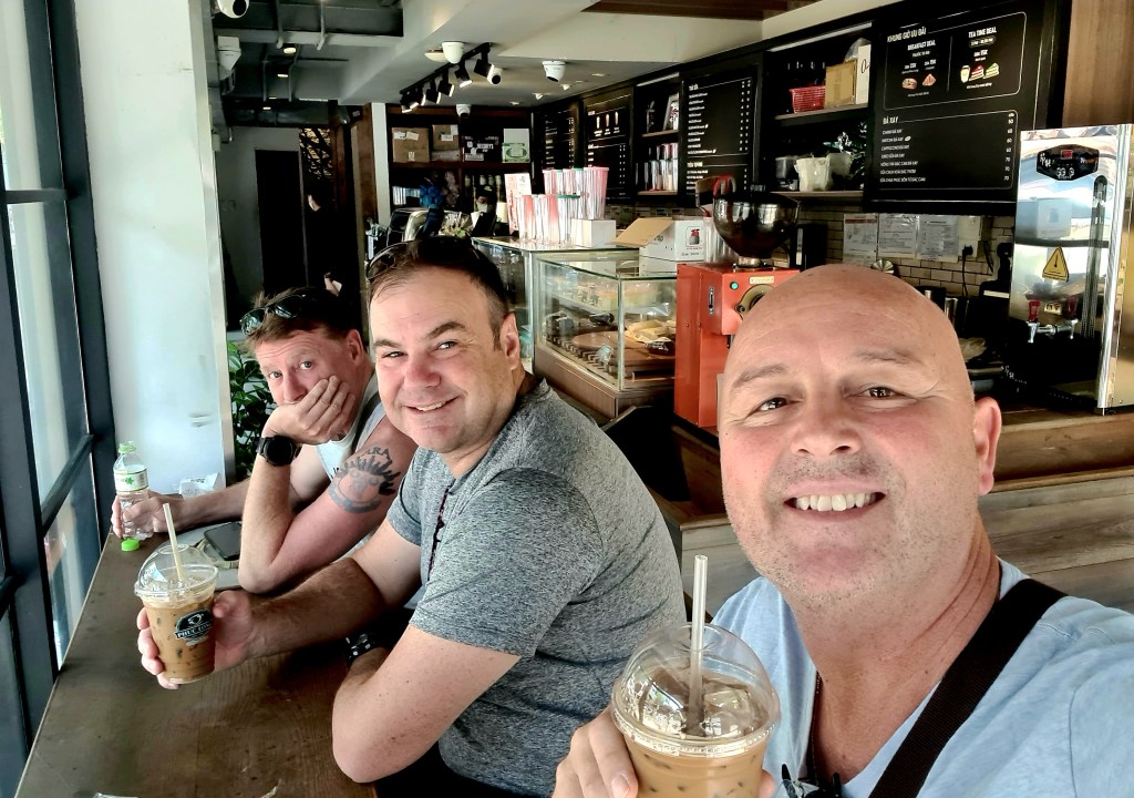 Three men sitting at a café table, smiling for the camera while holding iced drinks. The background features a coffee bar with a menu and various items on display.