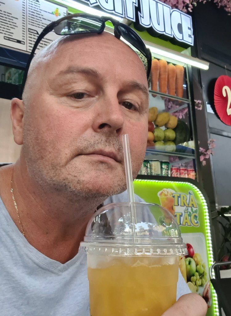 A man holding a cup of orange juice with a straw, wearing sunglasses, with a vibrant fruit juice stand in the background.