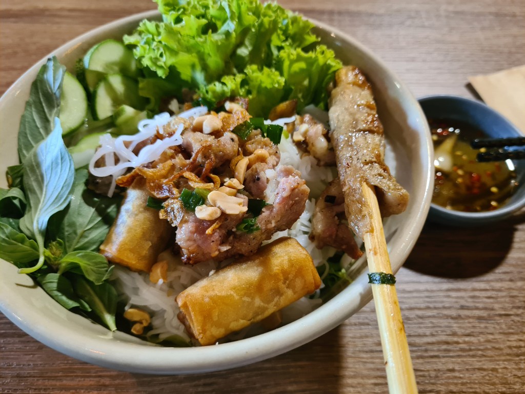A bowl of Vietnamese rice noodle dish featuring grilled meat, fresh herbs, lettuce, cucumber slices, fried spring rolls, and a dipping sauce on the side.
