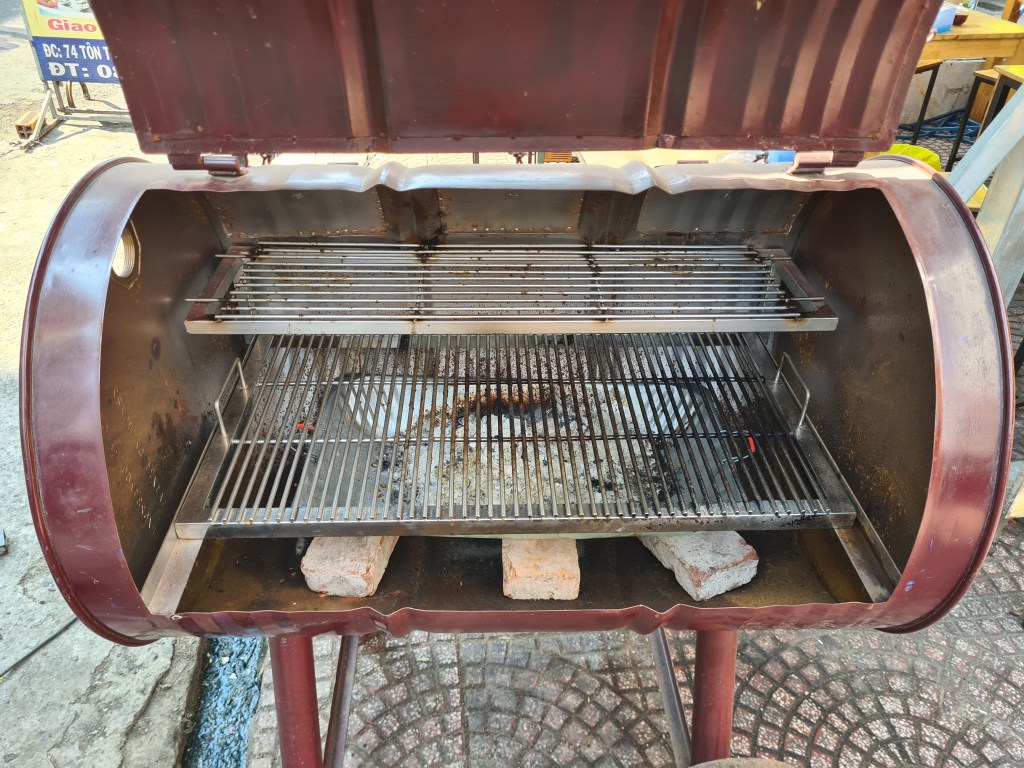 An open red charcoal grill with metal grates and a stone base, sitting on a paved outdoor surface.