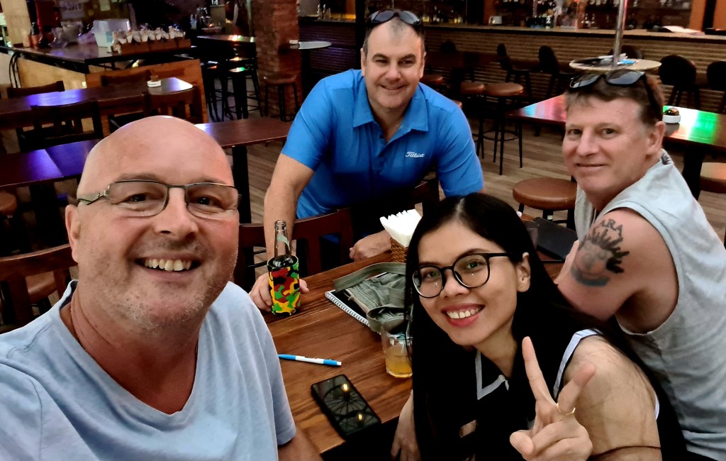 Group of four people sitting at a table in a casual dining setting, smiling at the camera. One person is holding a colorful drink, while another is making a peace sign.