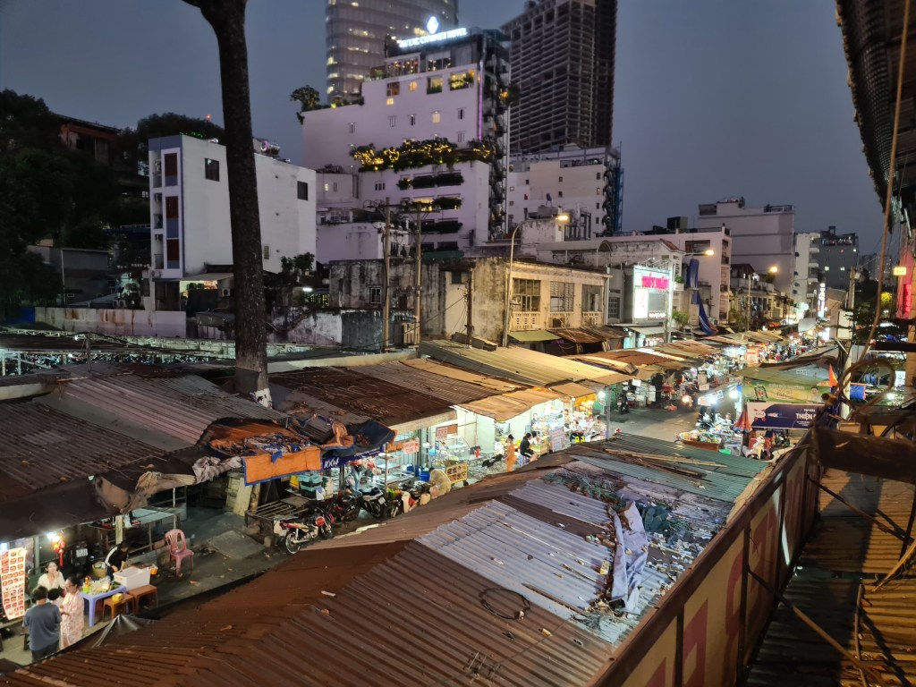 A bustling night market scene with rows of stalls and shops under metal roofs, illuminated by streetlights in an urban setting.