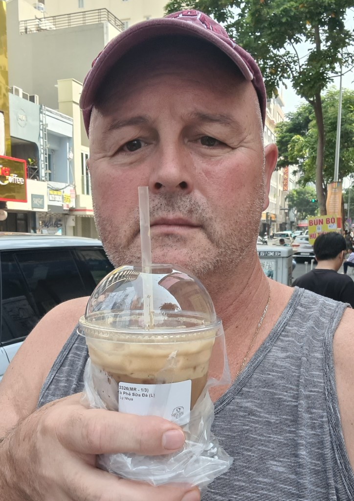 A man holding a plastic cup of iced coffee while standing on a city street, wearing a baseball cap and a sleeveless shirt.