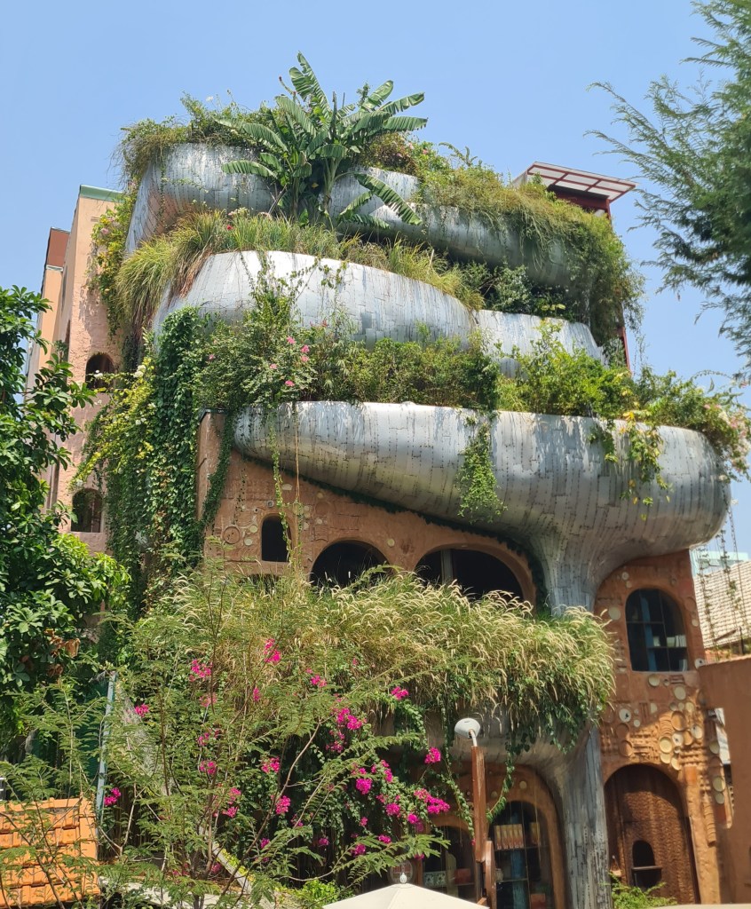 A unique, modern building with a wavy metallic facade, covered in lush greenery and flowers, set against a clear blue sky.