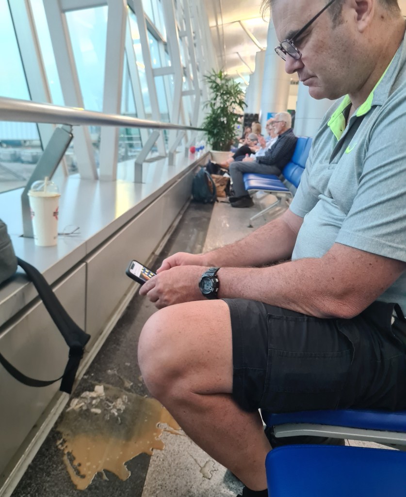 A man sitting in an airport terminal, focused on his phone, with spilled liquid on the floor beside him and other travelers in the background.