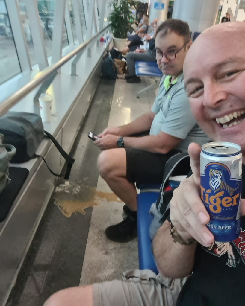 A man in a casual shirt smiles and holds a can of Tiger beer while taking a selfie in an airport seating area. Another man sits nearby looking at his phone, with a spill on the floor between them.