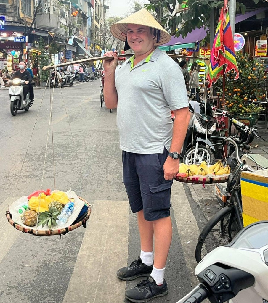 A man wearing a conical hat stands on a busy street, smiling while carrying a bamboo pole with two baskets of fruits, including bananas and pineapples.