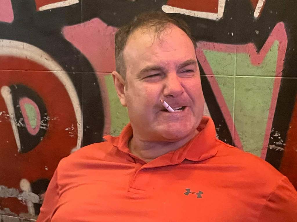 A man wearing a bright orange polo shirt is sitting against a colorful graffiti wall, smoking a cigarette and displaying an amused expression.