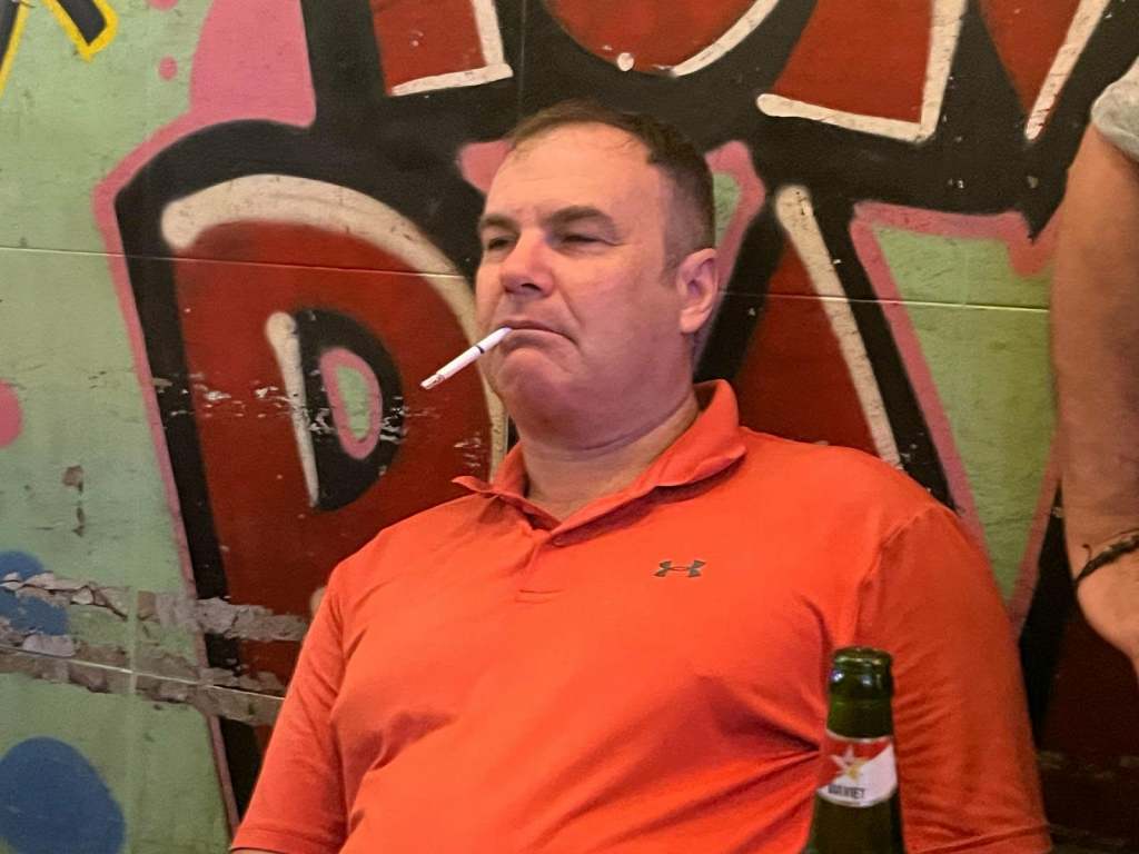 A man sitting casually with a cigarette in his mouth, wearing an orange polo shirt, against a colorful graffiti background.