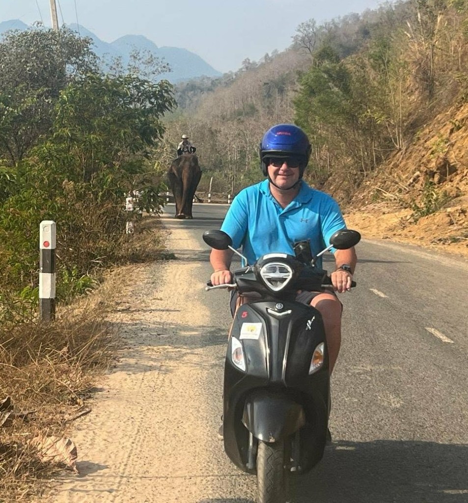 A person riding a scooter on a rural road, with an elephant and its rider visible in the background.