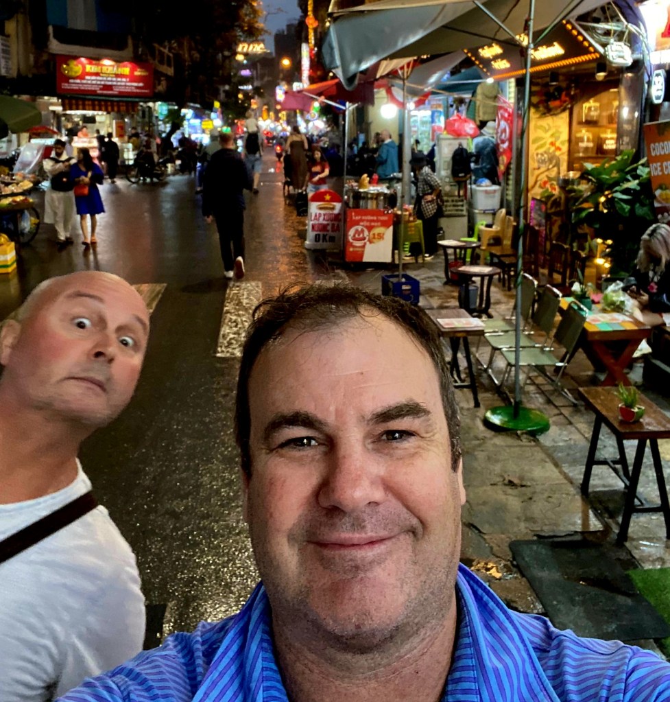 A selfie of two men in a busy street at night, with food stalls and shops lining the sides. One man is smiling at the camera while the other is making a funny face in the background.