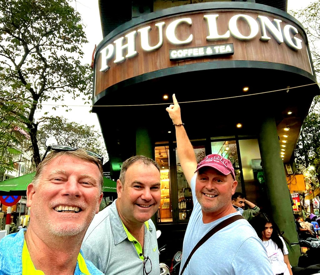 Three men smiling in front of the Phuc Long Coffee & Tea shop, with one man pointing at the sign.