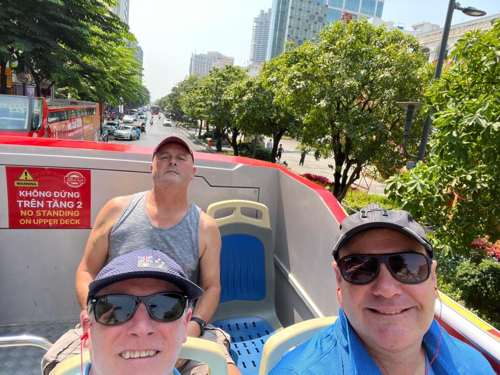 Three men enjoying a sightseeing bus ride on a sunny day, with one asleep and a city street and greenery in the background.